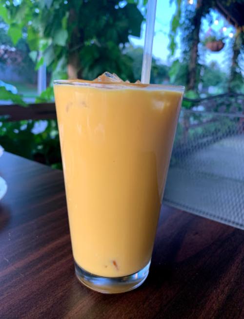 A tall glass of orange mango smoothie with ice, sitting on a wooden table surrounded by greenery.