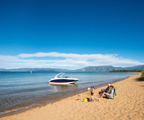 A sunny beach scene with a boat on the water and people relaxing on the sand. Mountains are visible in the background.