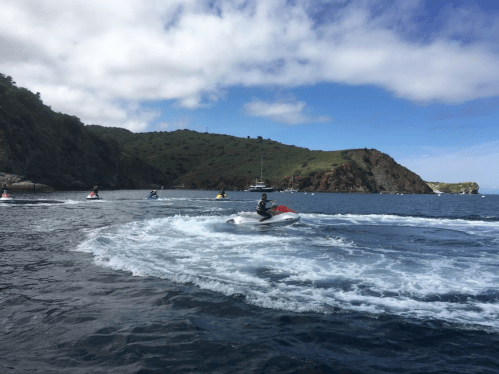 A group of jet skiers navigates through the ocean near a hilly coastline under a partly cloudy sky.