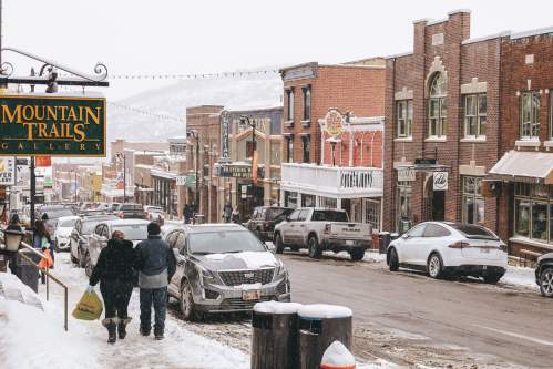 Snowy street scene with shops and parked cars, featuring people walking along the sidewalk in a winter setting.