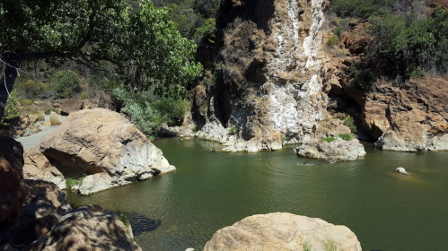 A serene natural landscape featuring a calm green pond surrounded by rocky cliffs and lush greenery.