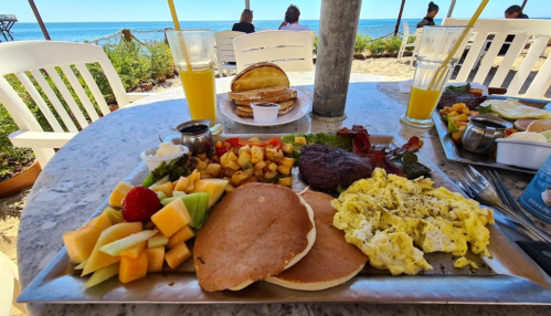 A beachside table with a hearty breakfast spread: pancakes, eggs, bacon, fruit, and drinks, with ocean views in the background.