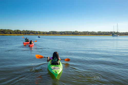 Two kayakers paddle on a calm river under a clear blue sky, with a sailboat in the background.
