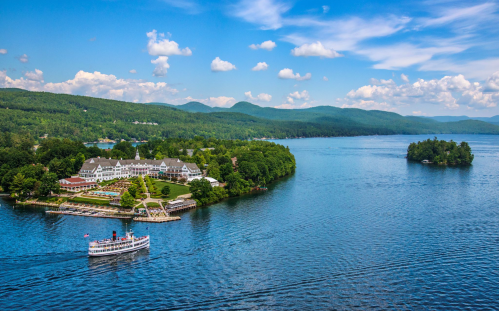A scenic view of a lake with a boat, surrounded by lush green hills and a resort along the shoreline under a blue sky.