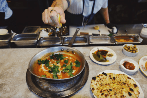 A chef adds a squeeze of lemon to a pan of curry, surrounded by various side dishes and naan bread.