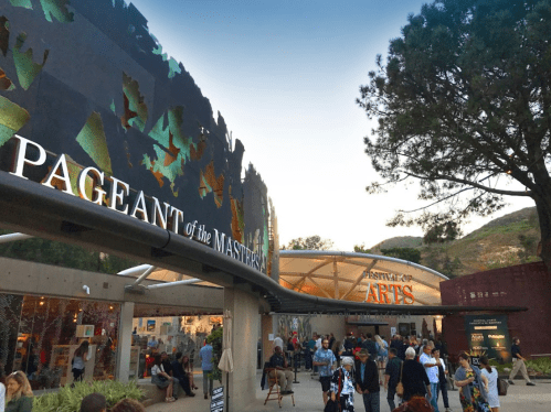 Crowd gathered outside the Pageant of the Masters venue, with art festival signage and trees in the background.