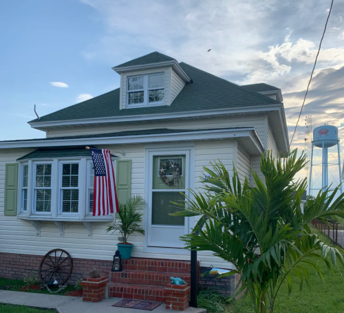 A charming white house with green shutters, an American flag, and a palm tree in front, under a cloudy sky.
