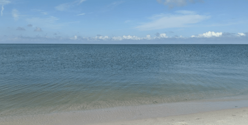 A serene beach scene with calm blue waters and a clear sky dotted with clouds.