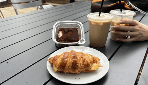 A croissant on a plate, a brownie in a container, and two iced drinks on a table.