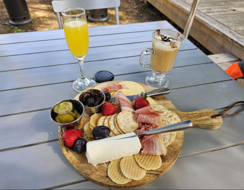 A wooden platter with cheese, crackers, fruits, and meats, accompanied by a mimosa and a coffee drink on a table.