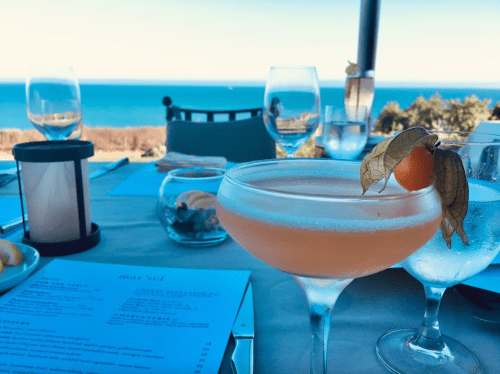 A cocktail with a garnish sits on a table overlooking a scenic ocean view, with menus and glasses in the background.