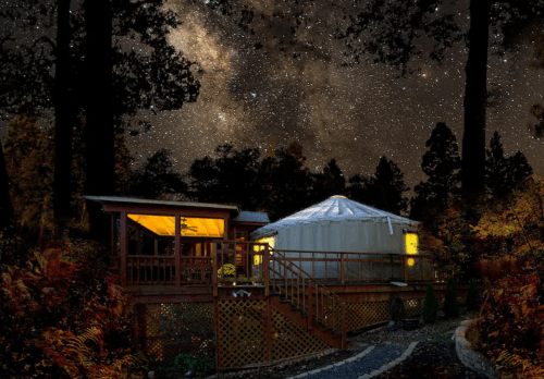 A cozy yurt and cabin under a starry night sky, surrounded by trees and illuminated windows.