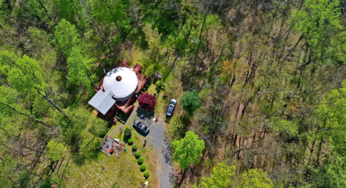 Aerial view of a circular building surrounded by trees, with a driveway and parked cars nearby.