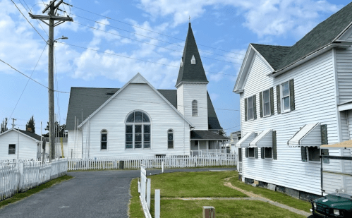 A white church with a tall steeple beside a white house, set against a blue sky with scattered clouds.