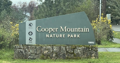 Sign for Cooper Mountain Nature Park, featuring park name and logo, surrounded by greenery and flowers.