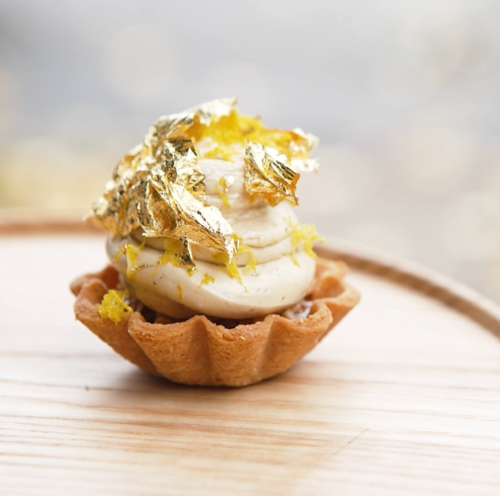 A small tart topped with creamy frosting and adorned with gold leaf, resting on a wooden surface.