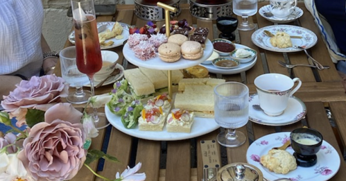 A table set with tea, pastries, and flowers, featuring a tiered stand of snacks and drinks in a cozy outdoor setting.