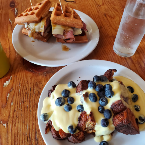 Two plates of food: one with waffle sandwiches and the other with bread pudding topped with blueberries and cream.