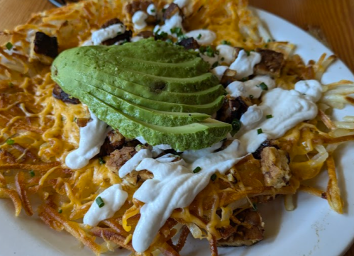 A plate of crispy hash browns topped with cheese, avocado slices, sour cream, and chopped herbs.