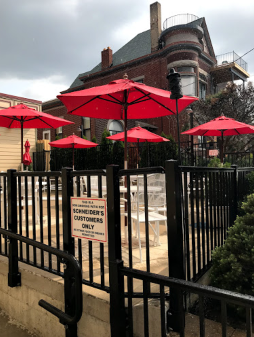 Outdoor seating area with red umbrellas, fenced off, next to a brick building. Sign reads "Schneider's Customers Only."