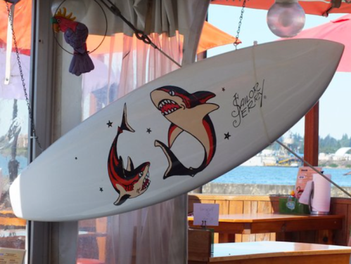 A white surfboard with colorful shark artwork hanging in a beachside setting.