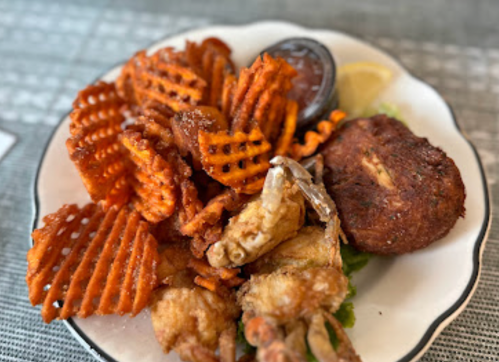 A plate of crispy fried seafood, including soft-shell crabs and a crab cake, served with waffle fries and dipping sauce.
