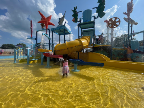A child plays in a colorful water park with a yellow pool and a large water slide under a sunny sky.