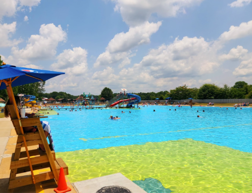 A sunny water park scene with a large pool, water slide, and people swimming under a blue sky with fluffy clouds.