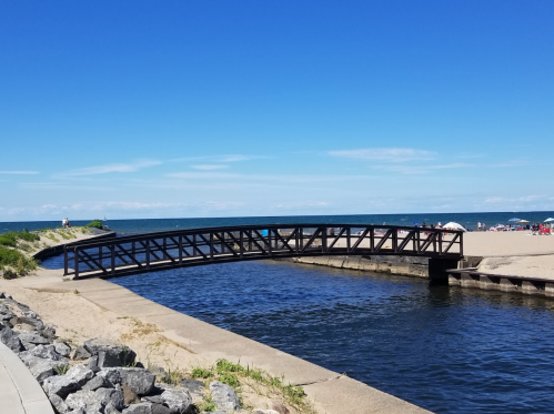 A black pedestrian bridge spans a calm waterway, leading to a sandy beach under a clear blue sky.
