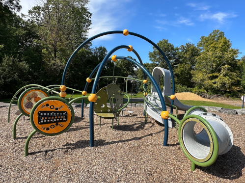 Colorful playground equipment with climbing structures and slides, surrounded by gravel and trees under a blue sky.