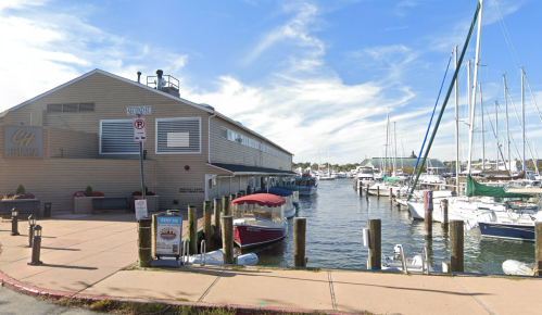 A marina with boats docked alongside a waterfront building under a blue sky with scattered clouds.