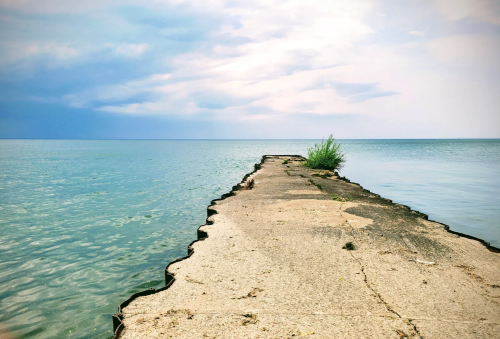 A weathered concrete pier extends into calm blue waters under a cloudy sky, with greenery at the edge.