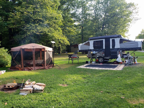 A campsite featuring a pop-up camper, a screened tent, a fire pit, and a picnic area surrounded by trees.