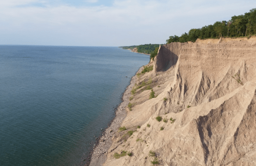 A scenic view of a sandy cliffside meeting a calm blue lake under a clear sky. Lush greenery lines the top of the cliff.