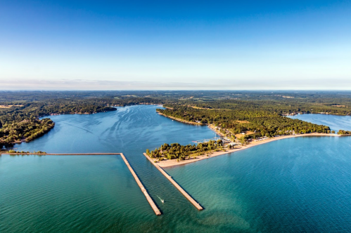 Aerial view of a serene lake with a sandy beach, piers, and lush green surroundings under a clear blue sky.