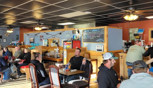 A cozy restaurant interior with patrons seated at tables, chatting, and enjoying their meals. A chalkboard menu is visible.