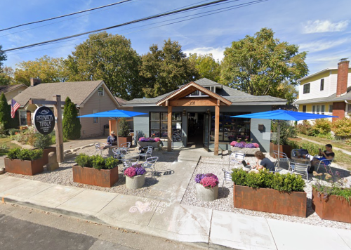 A cozy café with outdoor seating, blue umbrellas, and flower planters, surrounded by trees and residential buildings.