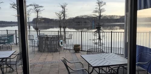 View of a calm lake with trees reflecting on the water, framed by outdoor seating and a railing.