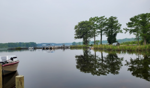 A calm lake scene with a boat docked, surrounded by trees and a misty sky, reflecting on the water's surface.