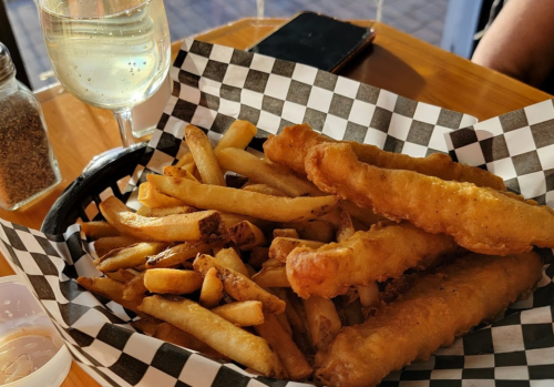 A basket of golden fried fish fillets and crispy French fries, with a glass of white wine in the background.