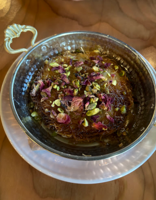 A silver dish filled with sweet, golden dessert topped with crushed pistachios and rose petals.