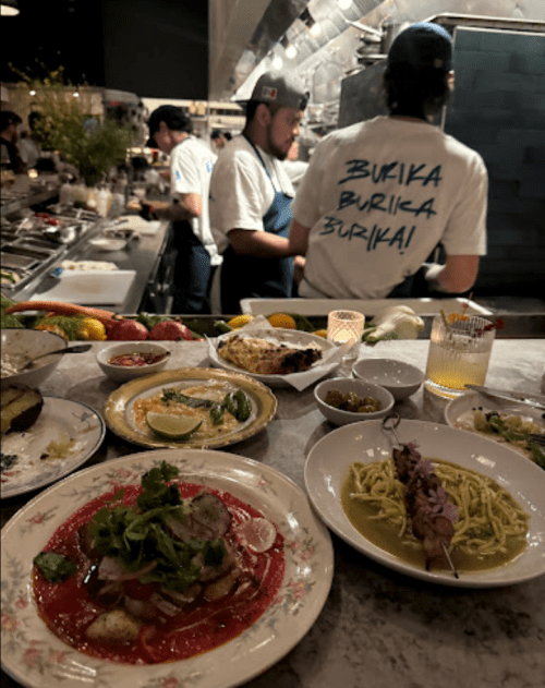 A vibrant restaurant scene with various dishes on a table and chefs working in the background.