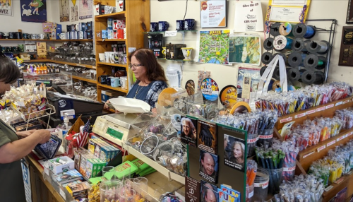 A cozy gift shop with colorful merchandise, a cashier, and a customer browsing items at the counter.