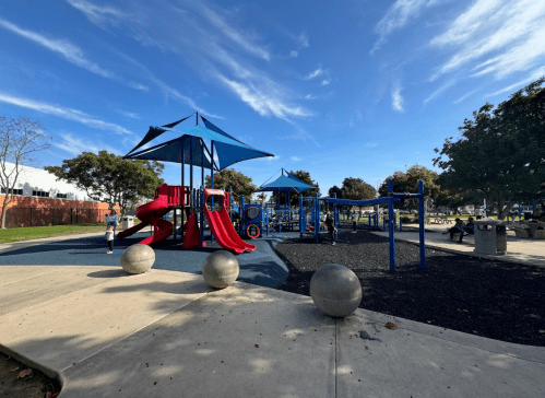 A colorful playground with slides and climbing structures under a blue sky, surrounded by trees and benches.
