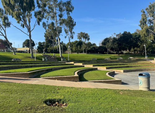A sunny park with curved seating areas, green grass, and trees under a clear blue sky.