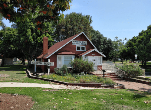 A red building with a sign reading "Manhattan Beach Historical Society," surrounded by greenery and trees.