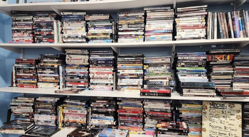 A cluttered shelf filled with various DVDs and CDs, with a sign listing prices for different media types.