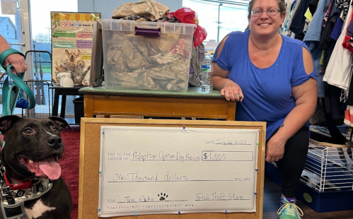 A woman smiles next to a dog and a large check for $1,000 made out to Adoption Option by House for pets.