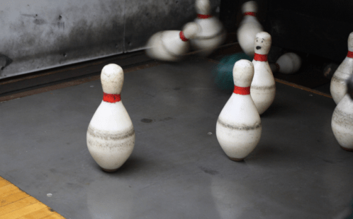 A bowling alley scene with several pins, one standing upright and others falling, with a bowling ball in motion.