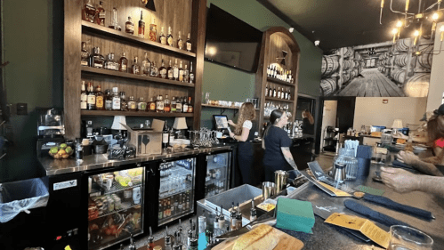 A bar with a wooden shelf displaying various liquor bottles, a bartender serving drinks, and patrons at the counter.